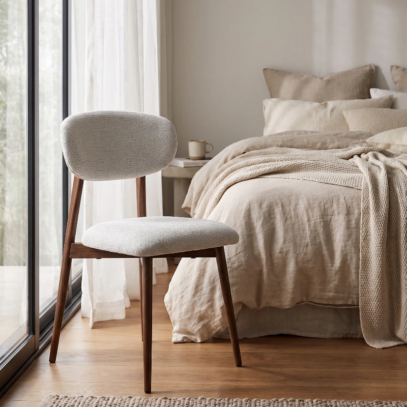 Light grey upholstered Japandi chair styled beside a bed in a neutral bedroom